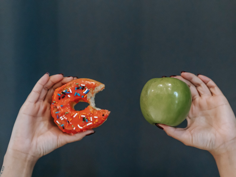 hands holding up a half eaten doughnut in the right hand and a green apple in the left hand