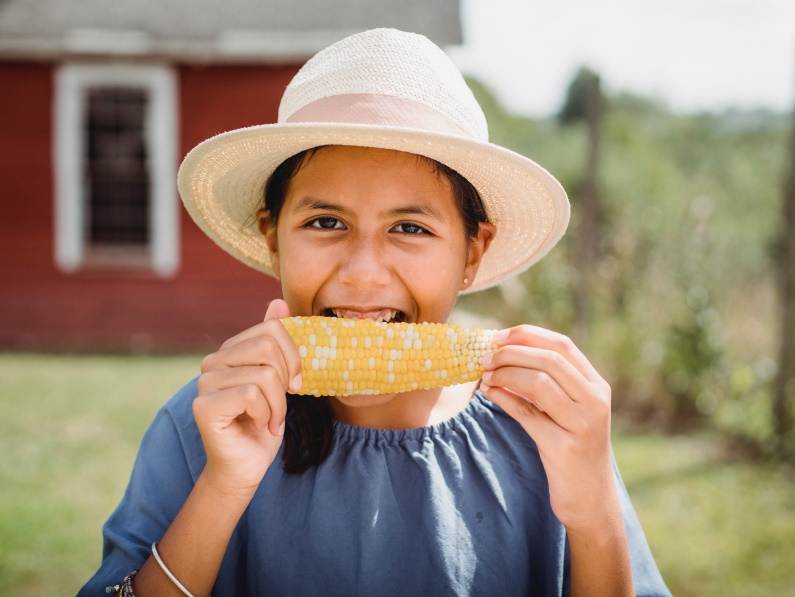 young girl with a sun hat on eating a cob of corn and smiling