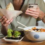 person eating a sandwich and salad, holding their phone